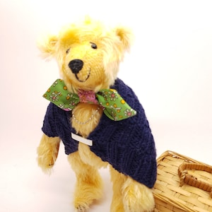 Könnte beinhalten: Ein gelber Teddybär, der einen blauen Strickpullover und eine grün-rosa florale Schleife trägt. Der Bär steht neben einem Weidenkorb.