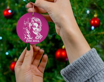 May include: A round, magenta Christmas ornament featuring a white portrait and the text "Ella 2024." The ornament is held by a person in front of a Christmas tree with red ornaments and green needles. A gold string is attached to the ornament.