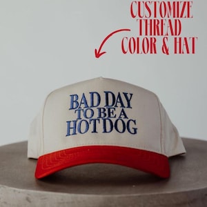 May include: A beige baseball cap with a red peak. The cap has the embroidered phrase "BAD DAY TO BE A HOT DOG" in blue. The text "CUSTOMIZE THREAD COLOR & HAT" is also visible.