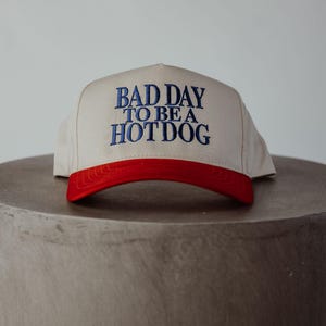 May include: A beige and red baseball cap with the text "Bad Day To Be A Hotdog" embroidered in blue.