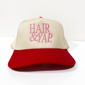 May include: A beige and red baseball cap featuring the pink embroidered text "HAIR & YAP". The cap has a red peak and a beige crown. This classic style hat is suitable for casual wear and is a popular accessory.