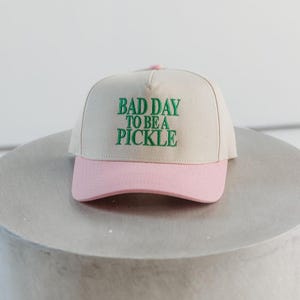 May include: A beige baseball cap with a pink brim. The cap features the green embroidered text, "BAD DAY TO BE A PICKLE." The hat is sitting on a gray surface.