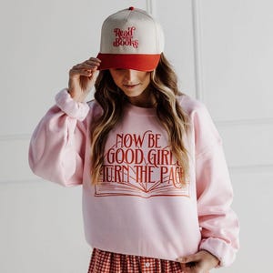 May include: A light pink sweatshirt with the text "NOW BE GOOD GIRL TURN THE PAGE" in orange. The image also shows a baseball cap with "Read More Books" in red and white, and red and white checked shorts.