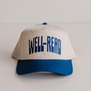 Puede incluir: Gorra de béisbol beige con visera azul y el texto "WELL-READ" en letras azules en negrita. La gorra tiene un diseño clásico de seis paneles con visera curva y botón en la parte superior.