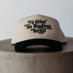 May include: A beige and black baseball cap with the text "I'd rather be reading smut" in black script. The cap is sitting on a gray, cylindrical surface.