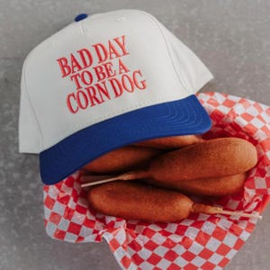 May include: A white baseball cap with a blue brim and the embroidered text "BAD DAY TO BE A CORN DOG" in red. The cap sits above a basket of corn dogs in a red and white checkered paper.