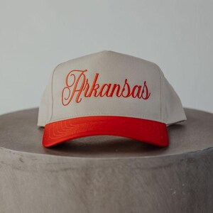 Puede incluir: Una gorra de béisbol beige con visera roja y la palabra "Arkansas" bordada en rojo en la parte delantera.