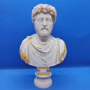 Puede incluir: Busto blanco de Marco Aurelio con rostro detallado, barba y cabello rizado. El busto tiene una túnica blanca con detalles dorados y está montado sobre un pedestal con el nombre "MARCUS AURELIUS" inscrito. La estatua está sobre un fondo azul.