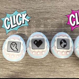 May include: Four pastel-coloured digital pet toys with pixelated icons of a ghost, heart, globe, and stars. The toys have a white border and are set against a wooden background. Comic book style speech bubbles with the word "CLICK" are above two of the toys.
