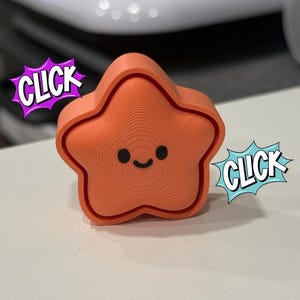 May include: An orange, star-shaped object with a smiling face. The object has a textured surface and is surrounded by two comic book style speech bubbles that say "CLICK".