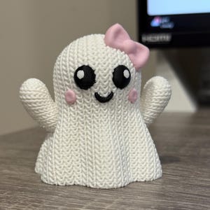 May include: A white ghost figurine with a knitted texture, featuring large black eyes, pink cheeks, and a small, smiling mouth. A pink bow adorns the top of the ghost's head. The figurine is a cute, whimsical decoration.