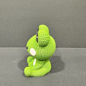 May include: A lime green bear figurine with a textured, knitted appearance. The bear has black eyes, a white spot on its cheek, and a white spot on its chest. The figurine is sitting on a gray surface.