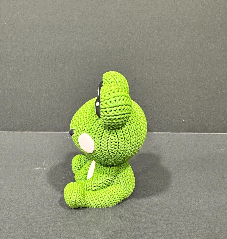 May include: A lime green bear figurine with a textured, knitted appearance. The bear has black eyes, a white spot on its cheek, and a white spot on its chest. The figurine is sitting on a gray surface.
