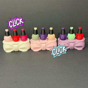 May include: A collection of miniature nail polish bottles in various colors, each with a heart-shaped top. The bottles are arranged in sets of three on bow-shaped holders. The word "CLICK" is displayed in a comic book style.