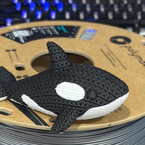 Puede incluir: Una figura de orca de impresión 3D en blanco y negro con una superficie texturizada. La figura está sentada sobre un carrete de filamento gris con el logotipo de Polymaker.