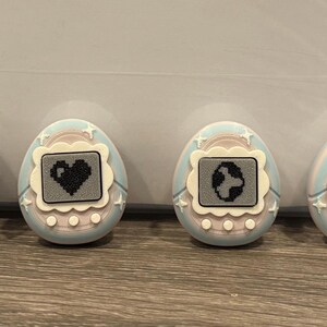 May include: Four pastel-coloured, egg-shaped digital pet toys. Each has a small screen displaying a pixelated graphic: a ghost, a heart, a globe, and two stars. The toys have a light blue and pink colour scheme with white accents.
