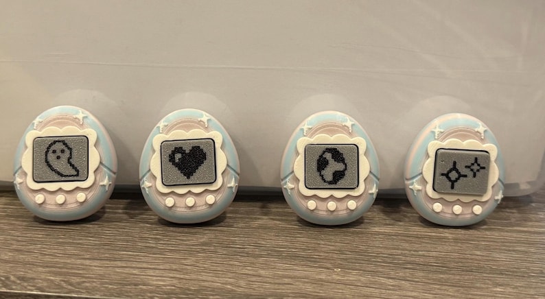 May include: Four pastel-coloured, egg-shaped digital pet toys. Each has a small screen displaying a pixelated graphic: a ghost, a heart, a globe, and two stars. The toys have a light blue and pink colour scheme with white accents.