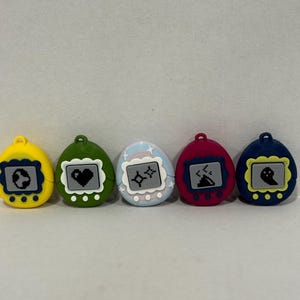 May include: Five colourful, egg-shaped digital pet toys. Each has a different colour shell: yellow, green, light blue, red, and dark blue. The screens display pixelated graphics of a globe, heart, astrological symbols, volcano, and ghost.