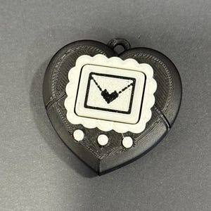 May include: A black and white heart-shaped pendant with a decorative border. The pendant features a square design with a heart and envelope illustration. Three white circles are below the square design.