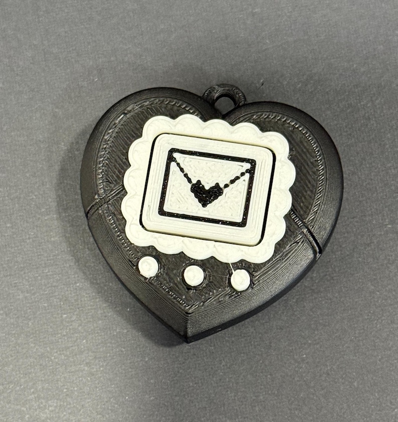 May include: A black and white heart-shaped pendant with a decorative border. The pendant features a square design with a heart and envelope illustration. Three white circles are below the square design.