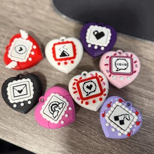 May include: A collection of heart-shaped accessories in various colors, including red, white, black, pink, and purple. Each accessory features a pixelated design within a white border. The accessories are arranged on a wooden surface.
