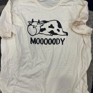 May include: Cream-colored t-shirt featuring a black and white cartoon cow graphic. The cow is lying down with the word "MOOOOODY" printed below in black block letters. The shirt has a crew neck and short sleeves.