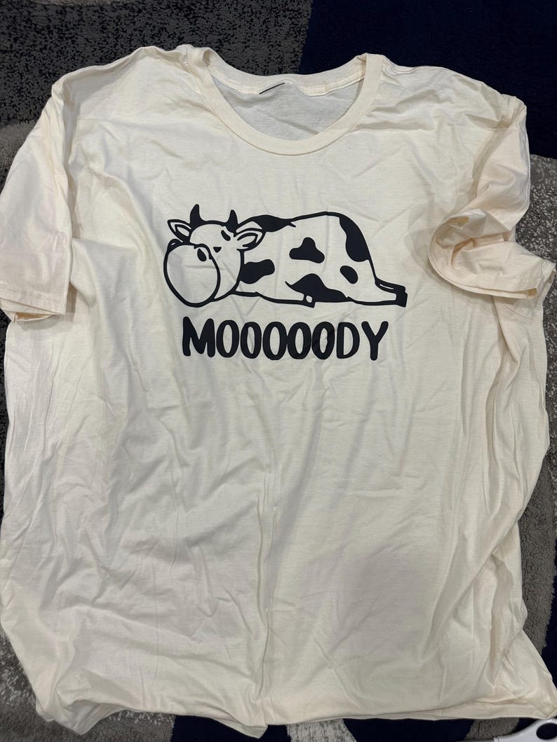 May include: Cream-colored t-shirt featuring a black and white cartoon cow graphic. The cow is lying down with the word "MOOOOODY" printed below in black block letters. The shirt has a crew neck and short sleeves.