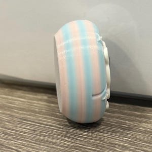 May include: A small, rounded object with vertical stripes of light blue and pink. The object has a white, decorative element on one side. The item appears to be 3D-printed and sits on a wooden surface.