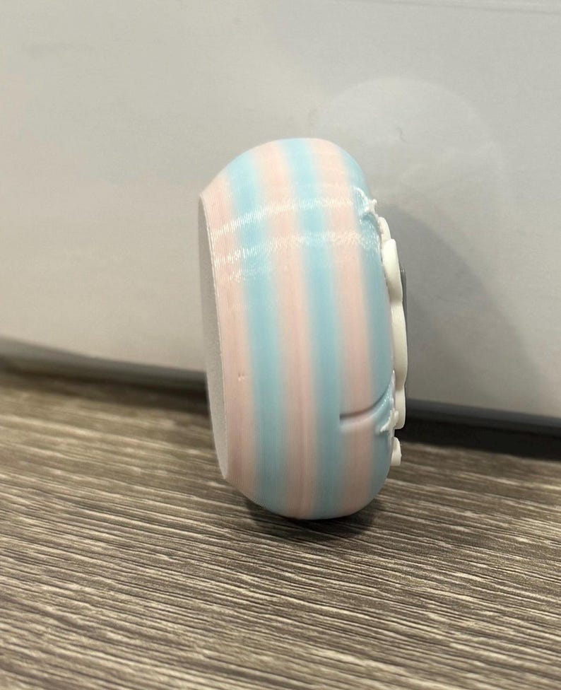 May include: A small, rounded object with vertical stripes of light blue and pink. The object has a white, decorative element on one side. The item appears to be 3D-printed and sits on a wooden surface.