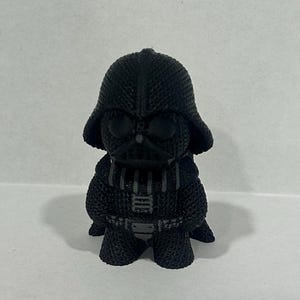 May include: A black and gray Darth Vader figurine with a textured surface. The figurine is standing on a white surface.