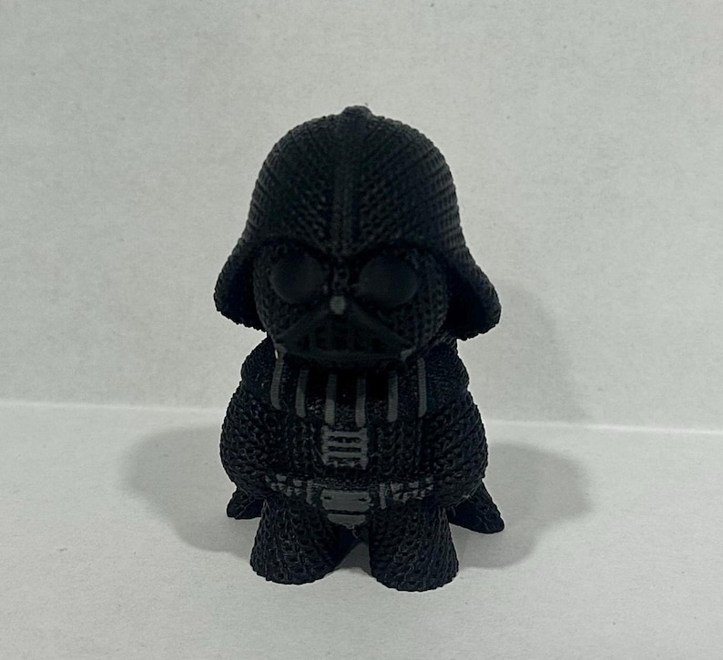 May include: A black and gray Darth Vader figurine with a textured surface. The figurine is standing on a white surface.