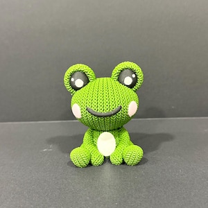 May include: A green knitted frog figurine with large black and white eyes, pink cheeks, and a smiling mouth. The frog has a white belly and is sitting on a dark surface. The frog is a cute, whimsical decorative item.