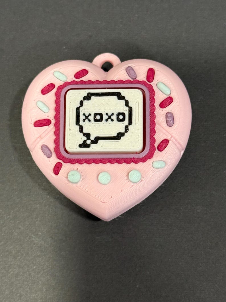 May include: A pink heart-shaped pendant with a rectangular center displaying the text "XOXO" in a speech bubble. The pendant is decorated with sprinkles and small circles.
