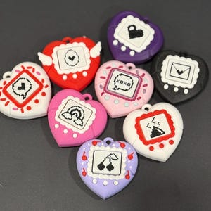 May include: A collection of heart-shaped accessories in various colors, each featuring a small screen with different pixelated icons. Colors include red, pink, purple, white, and black. Some have decorative elements like wings or cherries.