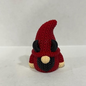 May include: A red gnome figurine with black horns and a black beard. The gnome's hat and body are knitted, with tan accents on the beard and feet. The figurine is a whimsical decorative item.