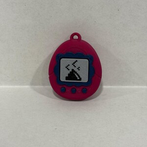 May include: A pink digital pet device with a blue border around the screen. The screen shows a pixelated graphic of a creature. The device has three buttons at the bottom and a loop at the top for attachment.
