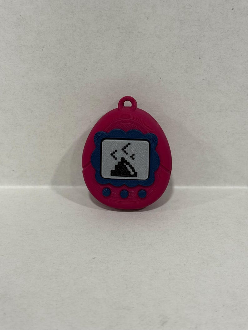 May include: A pink digital pet device with a blue border around the screen. The screen shows a pixelated graphic of a creature. The device has three buttons at the bottom and a loop at the top for attachment.