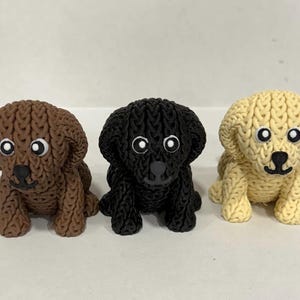 May include: Three small, knitted dog figurines in a row. The dogs are brown, black, and yellow, each with white eyes and a black nose. The figurines are sitting down, with their front paws visible.