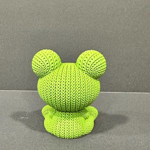 May include: A lime green frog figurine with a textured, knitted appearance. The frog has large, round eyes and a small, rounded body. The figurine is a decorative item.