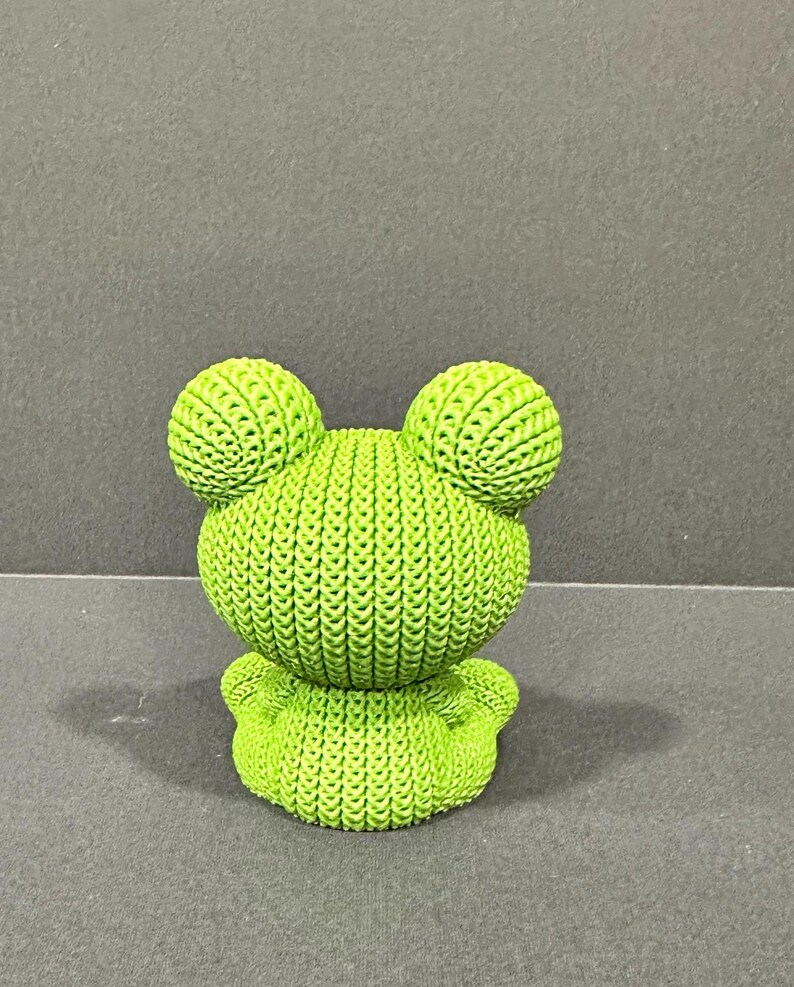May include: A lime green frog figurine with a textured, knitted appearance. The frog has large, round eyes and a small, rounded body. The figurine is a decorative item.