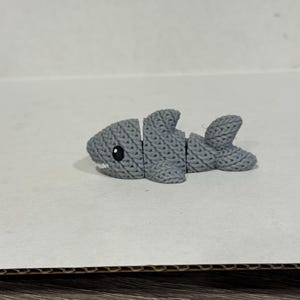 May include: A gray shark figurine with a textured, knitted appearance. The shark has a white tooth and a black eye. The body is segmented, and the tail is split into three sections. The figurine is on a light-colored surface.