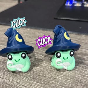 Op de afbeelding: Twee grillige kikkerbeeldjes, elk met een donkerblauwe tovenaars hoed met een gele halve maan. De kikkers zijn lichtgroen met witte buiken en grote zwarte ogen. Tekstballonnen boven de kikkers lezen "CLICK".