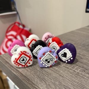 May include: A collection of heart-shaped trinkets in various colors, including white, pink, red, black, and purple. Each heart features a square design with a small graphic and decorative accents. The trinkets are arranged on a wooden surface.