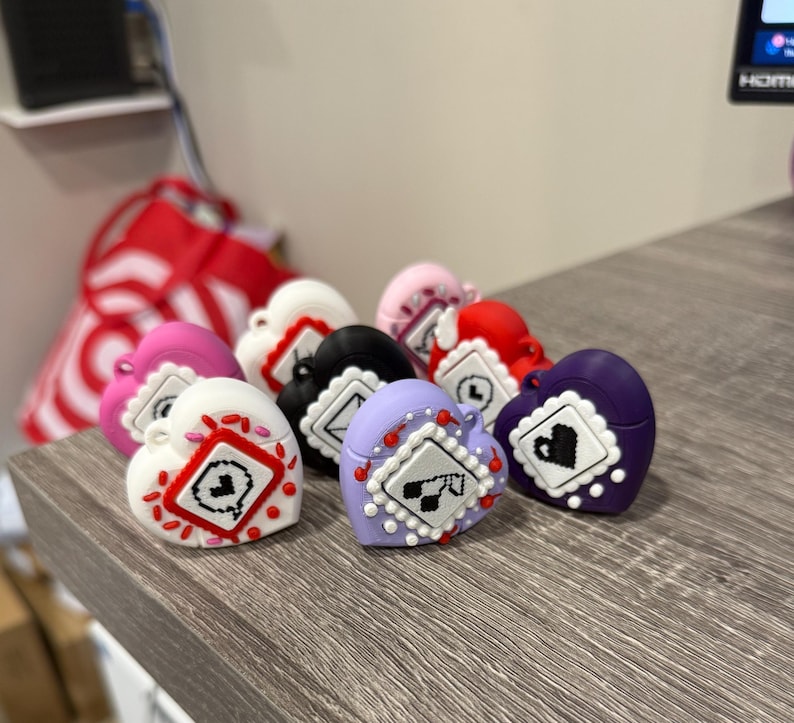 May include: A collection of heart-shaped trinkets in various colors, including white, pink, red, black, and purple. Each heart features a square design with a small graphic and decorative accents. The trinkets are arranged on a wooden surface.