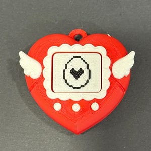 May include: A red heart-shaped pendant with white wings and a small screen. The screen displays a black heart icon within a white frame. The pendant has a loop at the top for hanging.