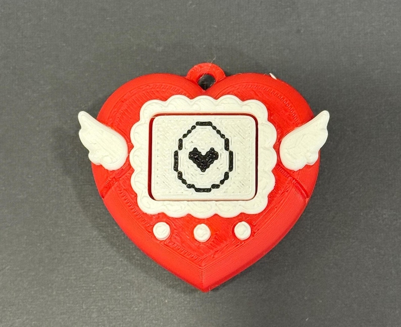 May include: A red heart-shaped pendant with white wings and a small screen. The screen displays a black heart icon within a white frame. The pendant has a loop at the top for hanging.