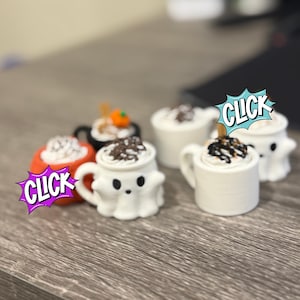 May include: A collection of small, white ghost-shaped mugs, each topped with a dollop of whipped cream and chocolate shavings. Some mugs have a small wooden stick. One mug is orange and another is black. The word "CLICK" is in a speech bubble.