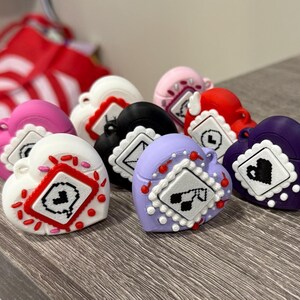May include: A collection of heart-shaped accessories in various colors, including white, pink, red, black, purple, and lavender. Each heart features a decorative square design with a unique graphic. The accessories are arranged on a wooden surface.