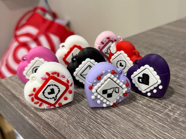 May include: A collection of heart-shaped accessories in various colors, including white, pink, red, black, purple, and lavender. Each heart features a decorative square design with a unique graphic. The accessories are arranged on a wooden surface.