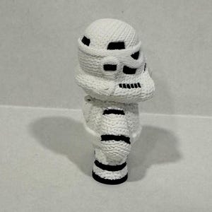 May include: White crocheted figurine of a Star Wars Stormtrooper with black details. The figurine is standing on a white surface.